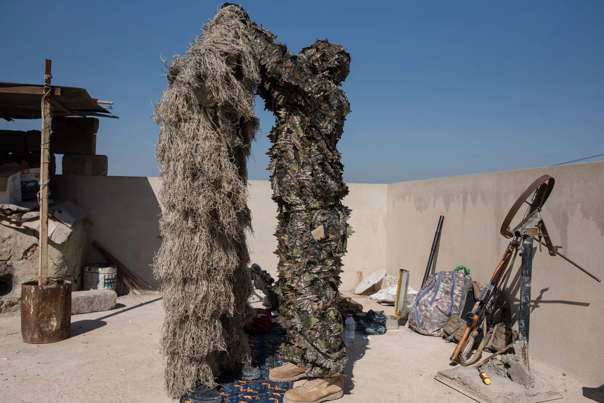 Two men in camouflage ghillie suits on a rooftop, one helping to adjust the other's suit
