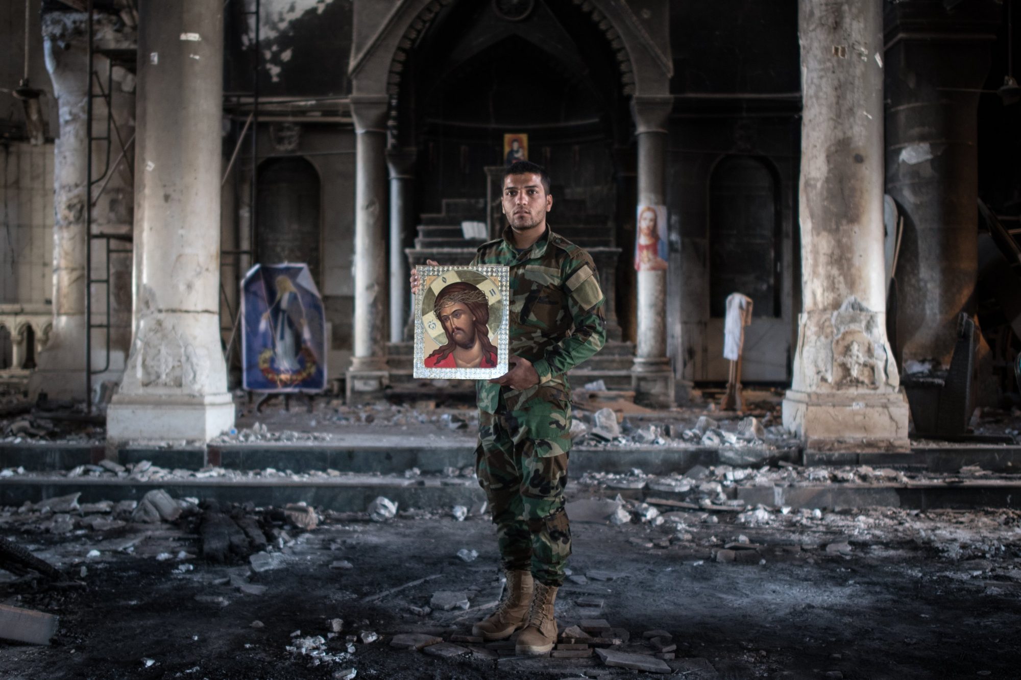 A man in camouflage holding an icon of Christ inside a burned-out church