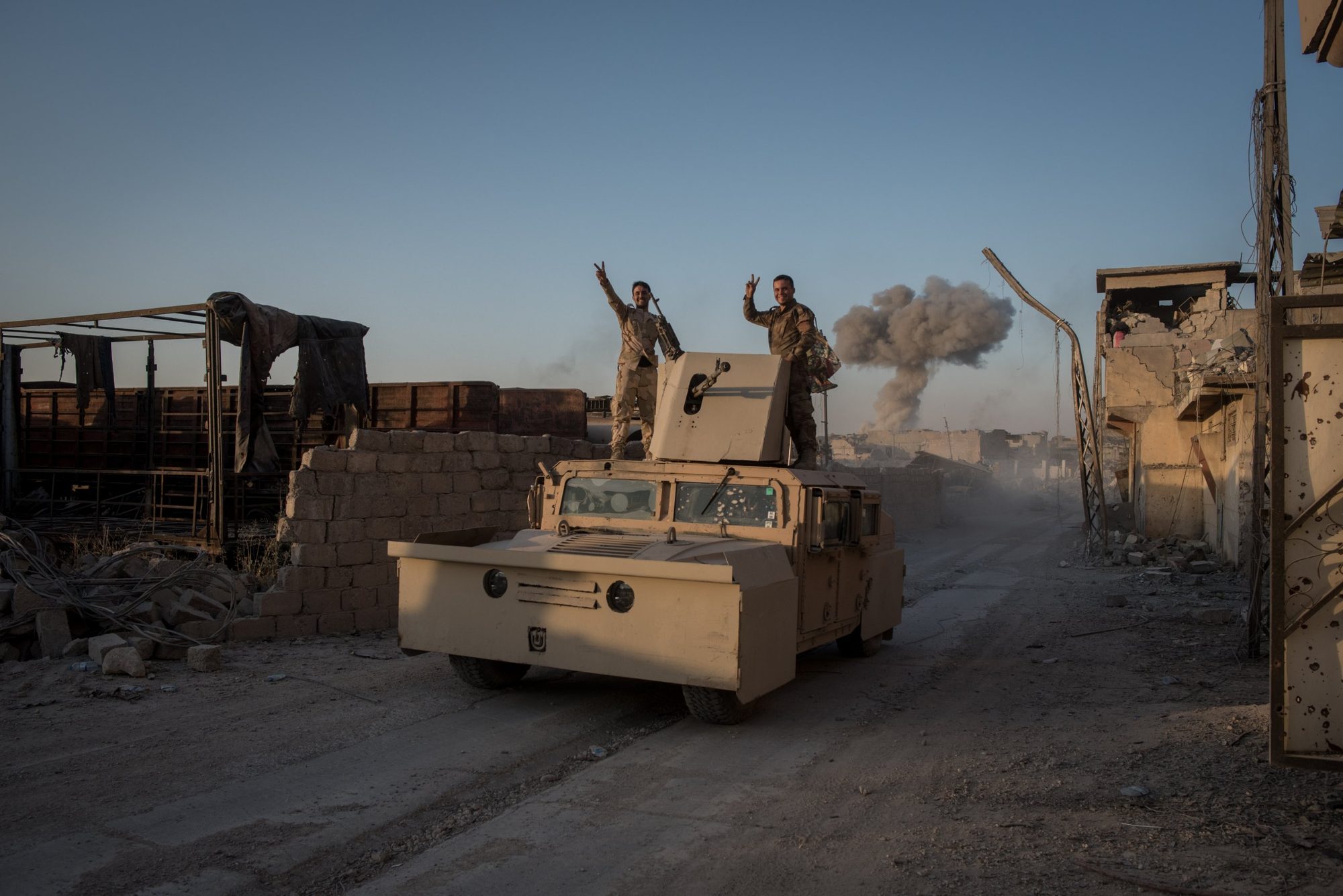 Two soldiers flashing peace signs from an armoured vehicle on a ruined street as smoke rises behind them